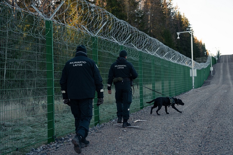 Kaksi rajavartijaa ja rajavartiokoira kulkee esteaidan viereisellä tiellä.
